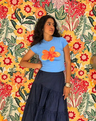 A woman wearing a light blue crewneck sunset tee with a large orange flower graphic on the front, paired with a dark navy blue tee.