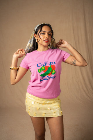 A woman modeling a pink graphic t-shirt with a green and red pepper illustration and the text 'My Gusta Caliente'