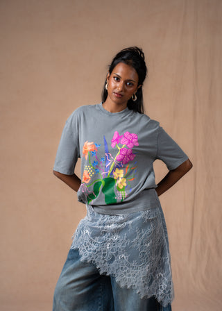 A model wearing a heather grey t-shirt with a colorful floral graphic print on the front, styled with a light blue lace wrap and blue jeans